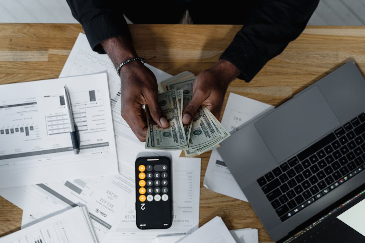 Finance staff counts cash after a fundraiser at a desk with a calculator, a laptop and income planning print outs